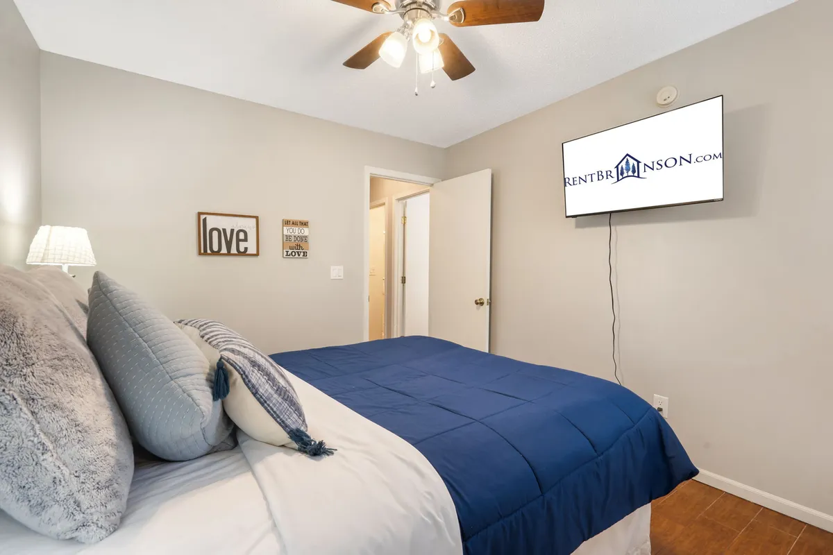 A warm, inviting bedroom with blue bedding, wall art that reads 'love,' and a wall-mounted television. The space exudes coziness and intimacy—perfect for couples or unwinding after a busy day.