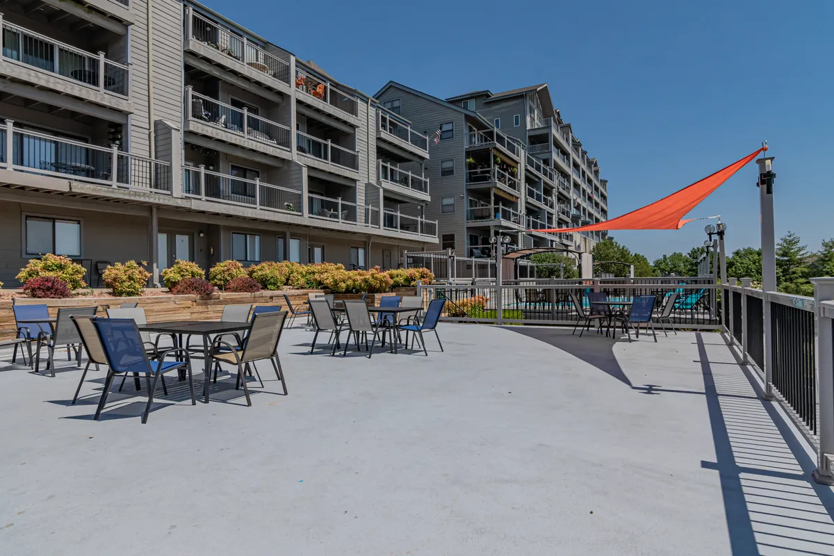 A casual outdoor seating area, surrounded by lush green plants and flowers, features tables under a shade sail. The scene suggests relaxed outdoor dining, socializing, and basking in the sun.