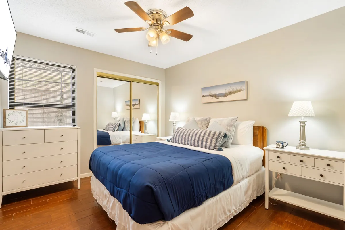 A cozy bedroom featuring a bed with a blue comforter, a mirrored wardrobe, and a nightstand with a lamp. The space is bathed in soft, natural light, creating a calm and restful ambiance that invites relaxation and restful sleep.