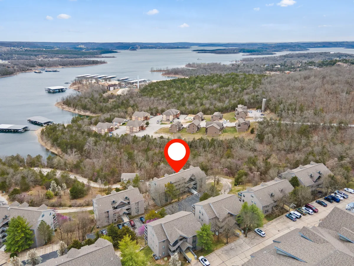 This image showcases the picturesque lakefront with Table Rock Lake in the foreground, surrounded by lush trees and residential buildings. The parking area is visible, emphasizing easy access for guests bringing boats or trailers. The tranquil water, combined with the nearby homes, underscores the seamless connection between outdoor adventure and comfortable living.