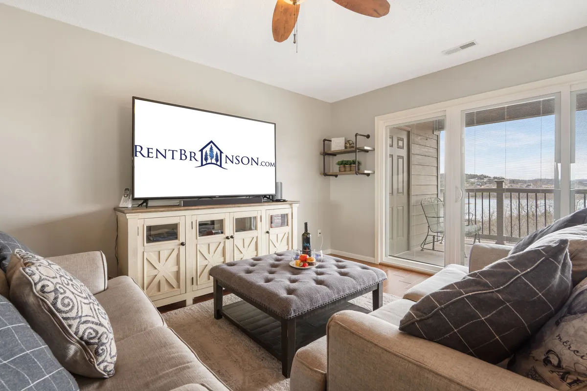 The image showcases a bright, inviting living room with a plush sofa, a sleek coffee table set for game nights, and a large television. Through sliding glass doors, a private balcony opens to sweeping views of Table Rock Lake, hinting at stunning sunsets and fresh lakeside breezes.