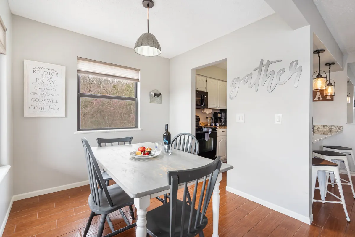 A warm and inviting dining area anchored by a large table, decorative wall art, and a window letting in natural light. The kitchen entrance is visible, creating a clear connection between cooking and dining spaces ideal for family meals and group conversations.